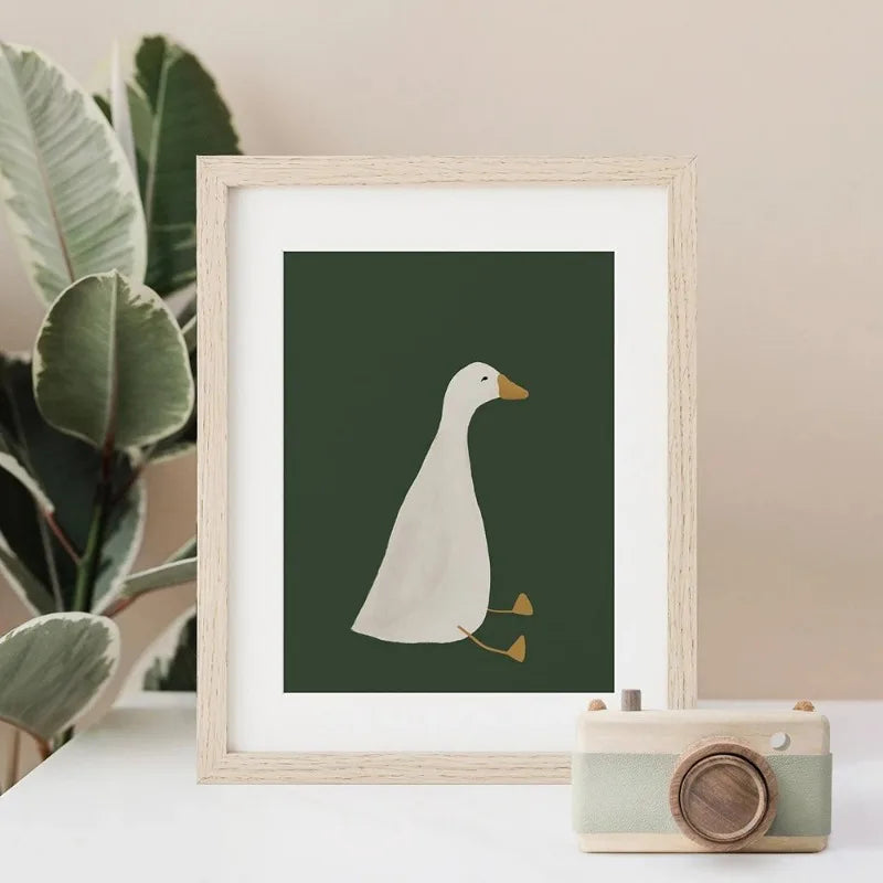 Framed artwork of a duck on a dark green background with a plant and camera in the background.