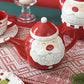 Red teapot shaped like a Santa Claus face on a table with Christmas decor.