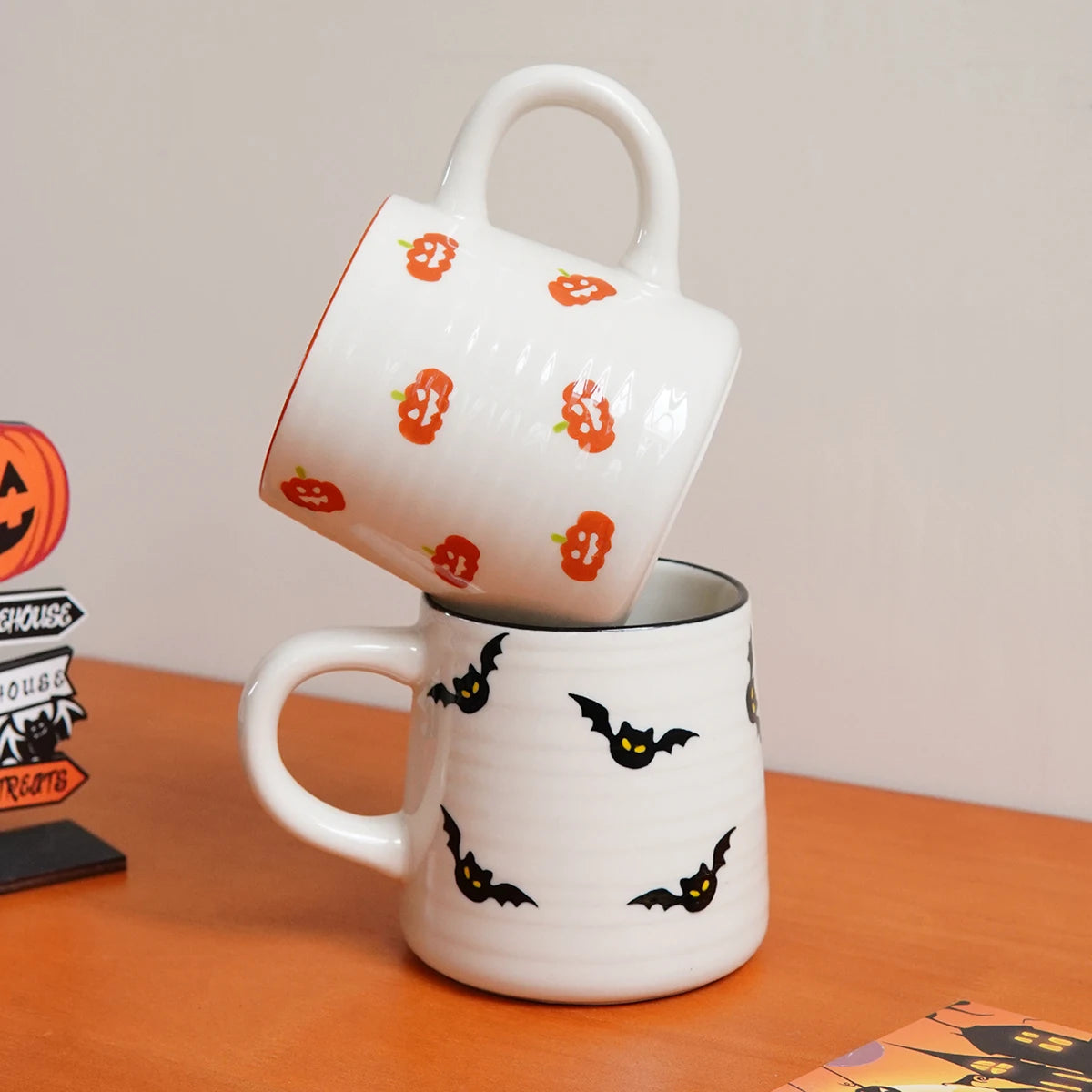 Two Halloween-themed mugs stacked on a wooden surface with pumpkins and bats designs.
