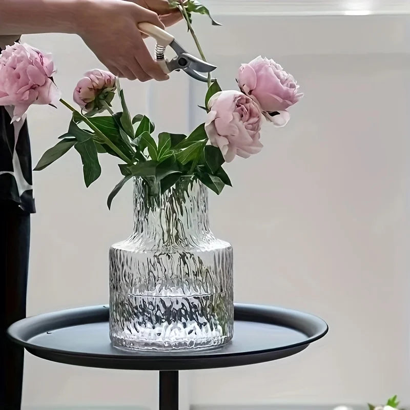 Person arranging pink flowers in a clear glass vase on a small round table.
