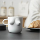White mug shaped like a breast on a kitchen counter with a plate of cookies and coffee maker in the background.