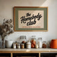 Framed poster with 'the Homebody Club' text on a kitchen counter with jars and decor.