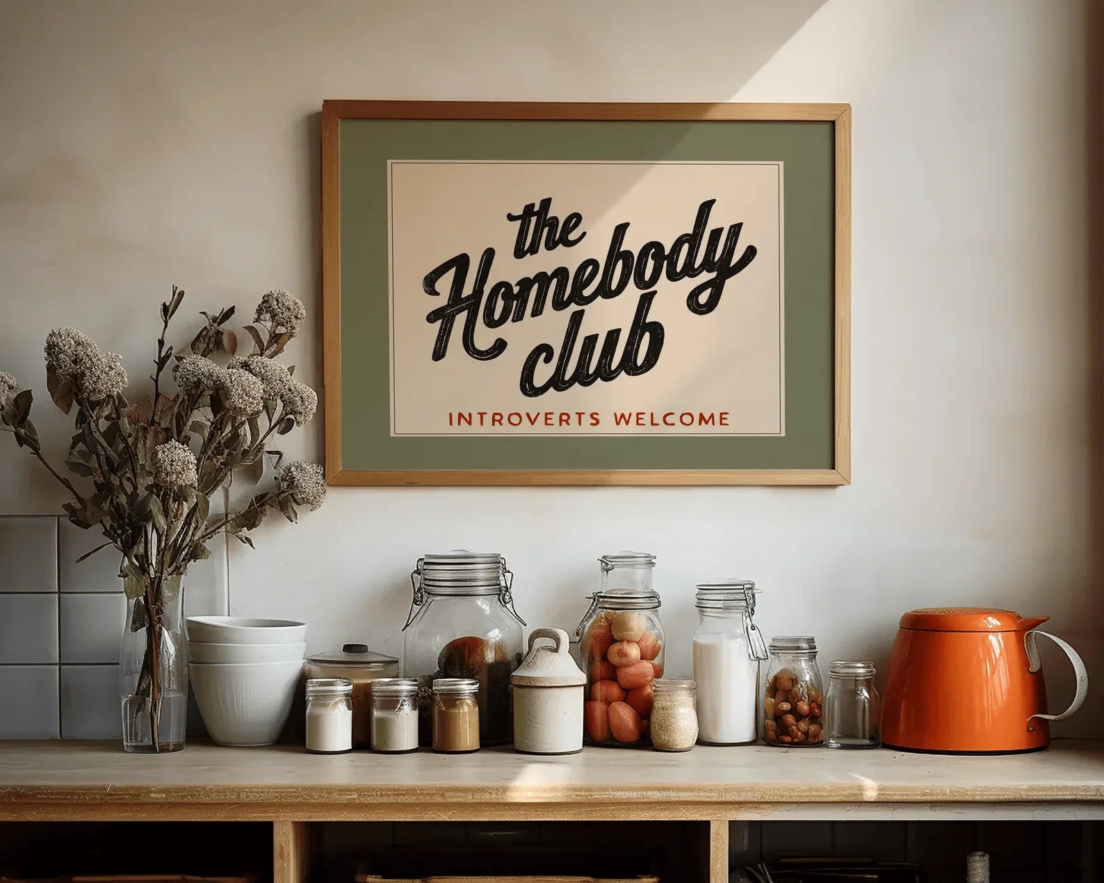 Framed poster with 'the Homebody Club' text on a kitchen counter with jars and decor.