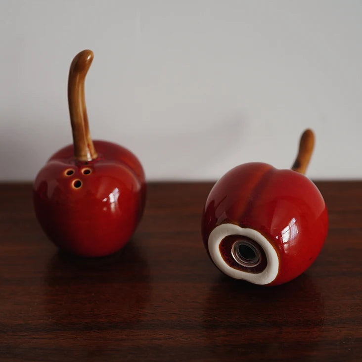 Two red ceramic cherries with brown stems on a wooden surface.