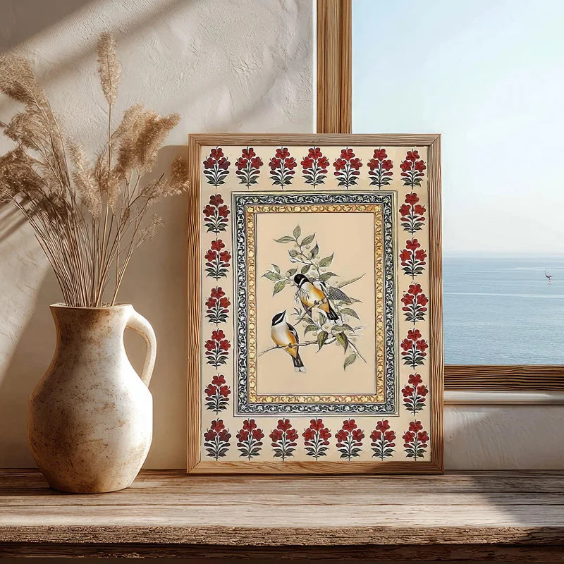 Framed artwork with floral and bird design next to a vase with dried plants on a wooden surface.