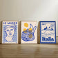 Three framed artworks on a wooden shelf against a beige wall.