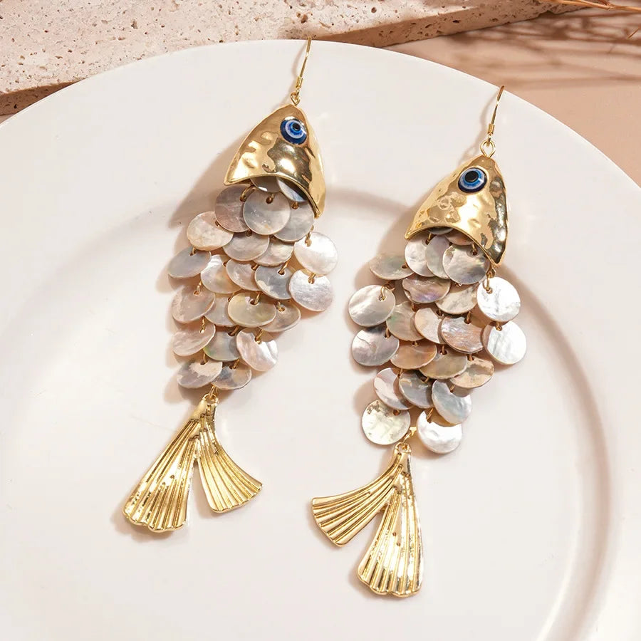 Gold fish-shaped earrings with shell details on a white plate.