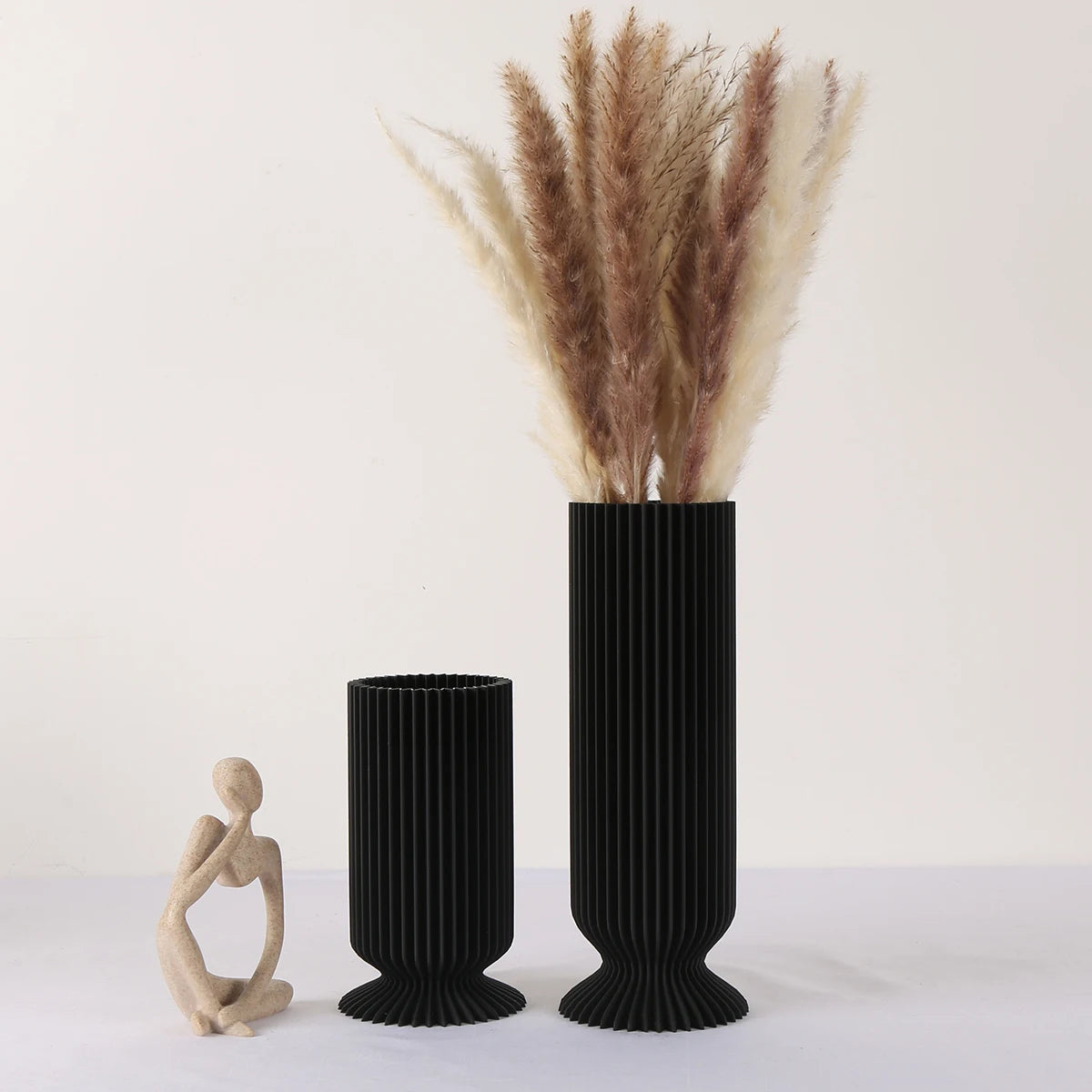 Two black textured vases with dried pampas grass and a small wooden sculpture on a white background