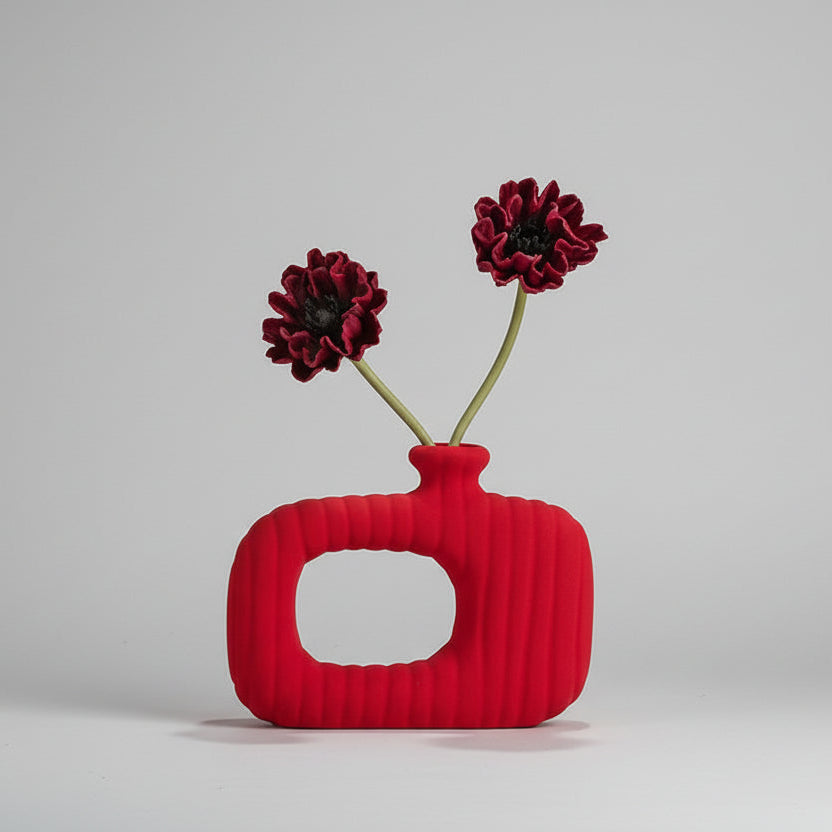 Red textured vase with flowers on a shelf against a neutral wall.