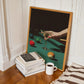 Framed artwork of a hand holding a cocktail glass on a green surface with books and a coffee cup below.