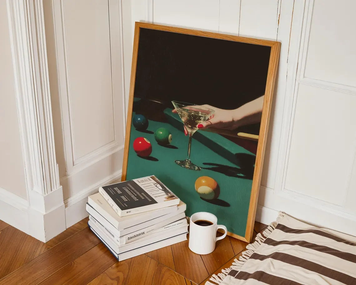 Framed artwork of a hand holding a cocktail glass on a green surface with books and a coffee cup below.