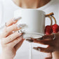 Person holding a white mug with a decorative handle featuring red cherries.