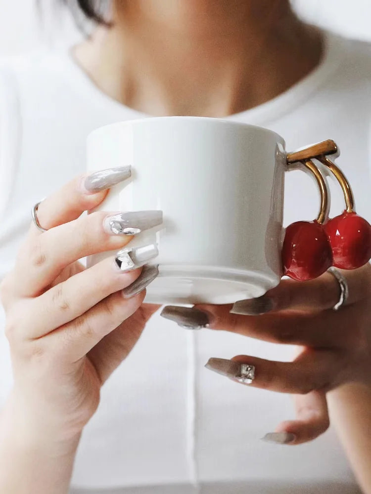 Person holding a white mug with a decorative handle featuring red cherries.