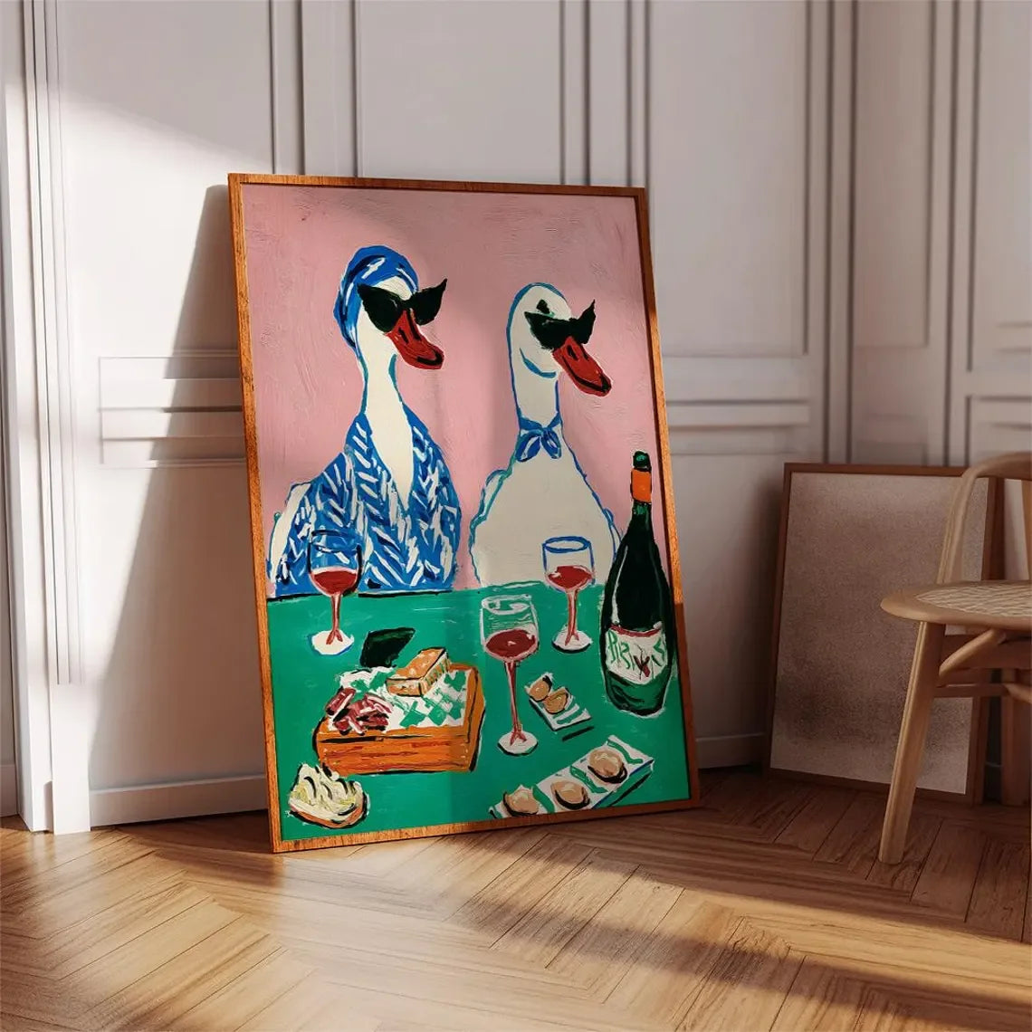 Framed artwork of two stylized ducks at a table with wine and food on a wall.