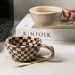 Person holding a ceramic mug with a checkered design on a desk next to a laptop and 'KINFOLK' book.