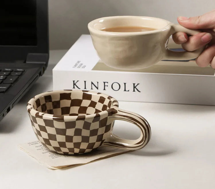 Person holding a ceramic mug with a checkered design on a desk next to a laptop and 'KINFOLK' book.