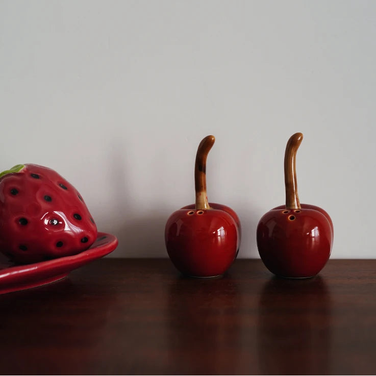 Ceramic strawberry and cherry salt and pepper shakers on a wooden surface with a plain background.