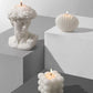 Three white candles, including a bust, shell, and bubble design, on a geometric gray surface.
