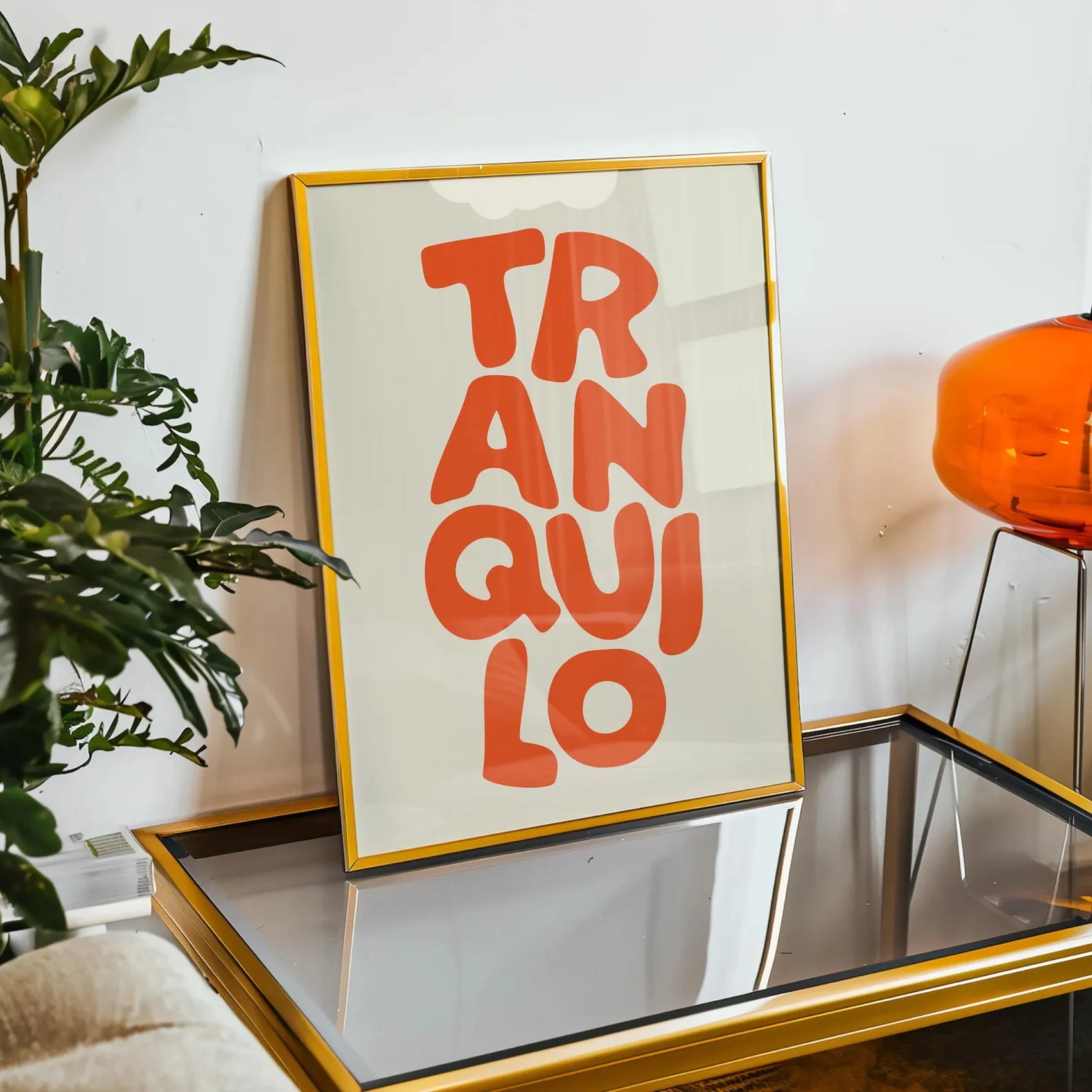 Framed artwork with 'TRANQUILO' text on a table with a plant and lamp in the background