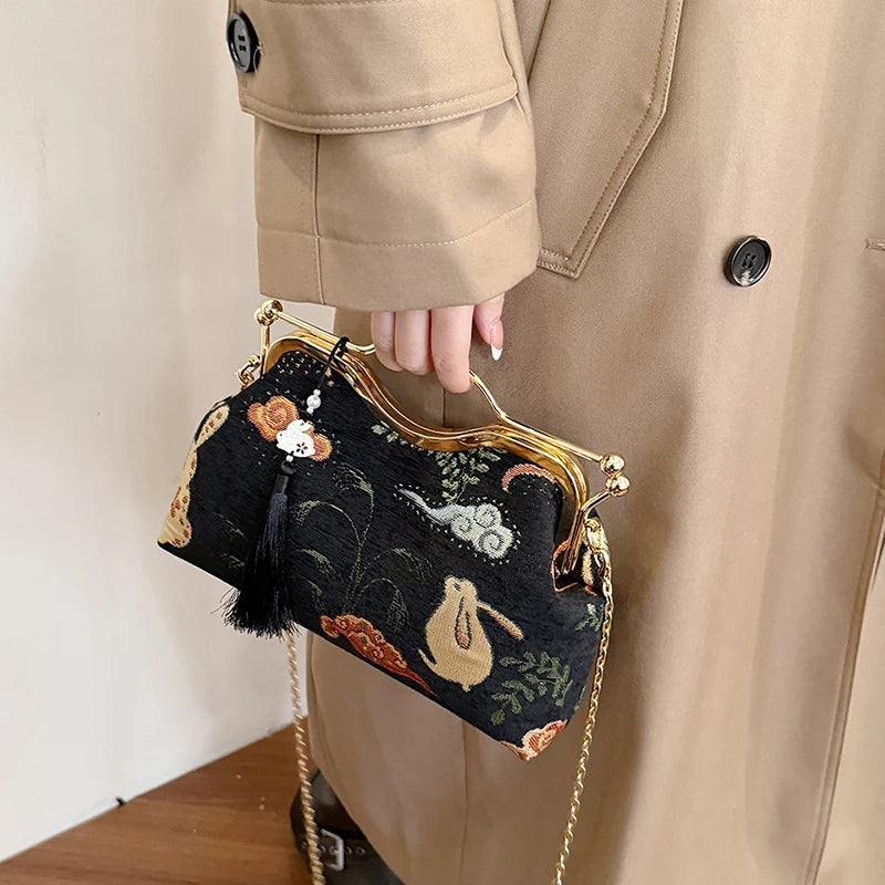 Person holding a black embroidered handbag with gold frame and chain strap, wearing a beige coat.