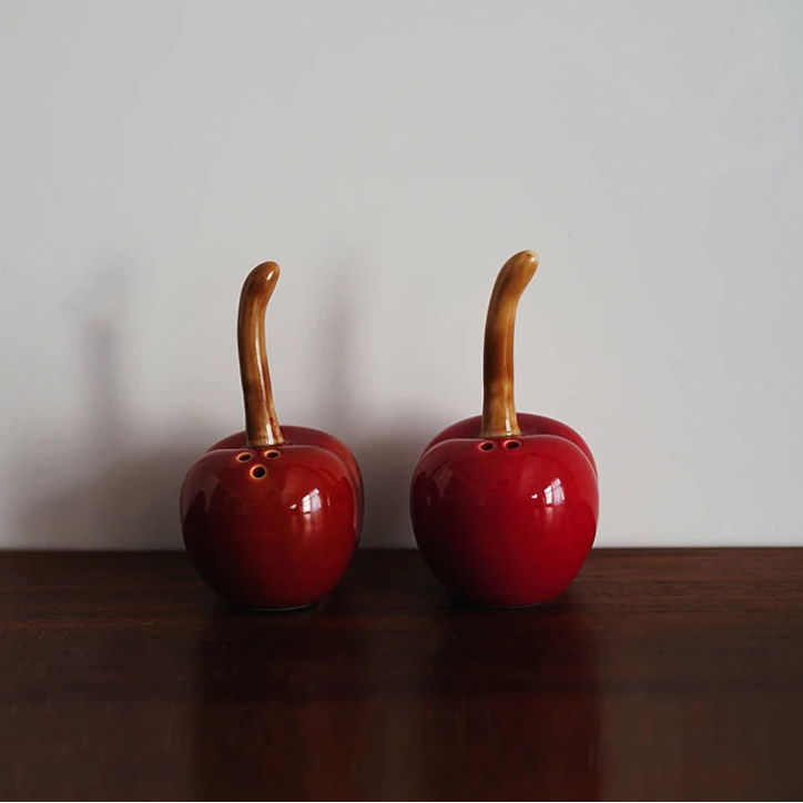 Two red ceramic apple-shaped items with wooden stems on a dark surface and light background.