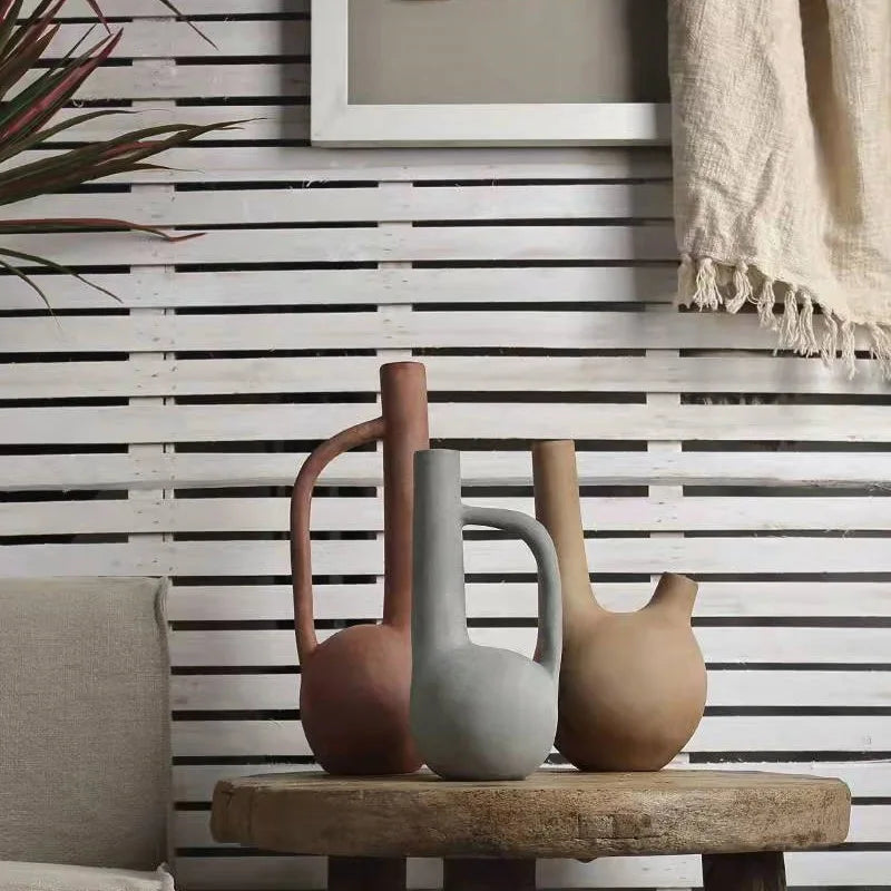 Three ceramic vases on a wooden stool against a slatted wall with a framed picture and blanket.