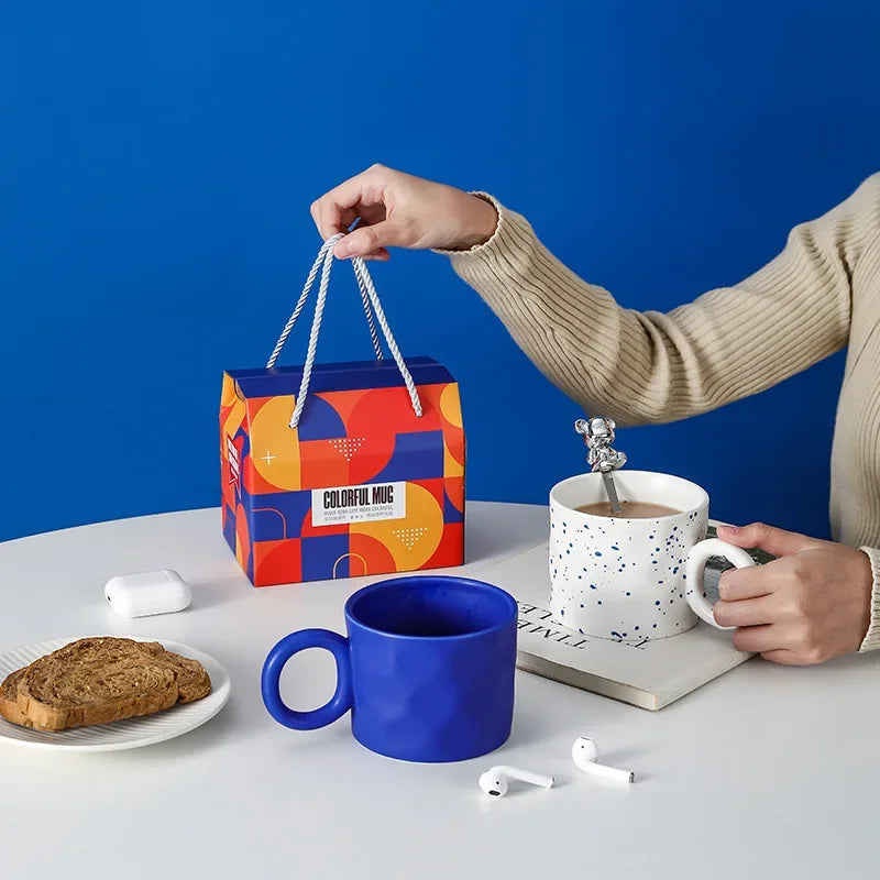Person holding a colorful gift bag with a blue mug, white cup, and toast on a table against a blue background.