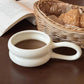 White ceramic mug with handle on a wooden surface, containing a dark liquid, next to an open book and a woven basket with bread.