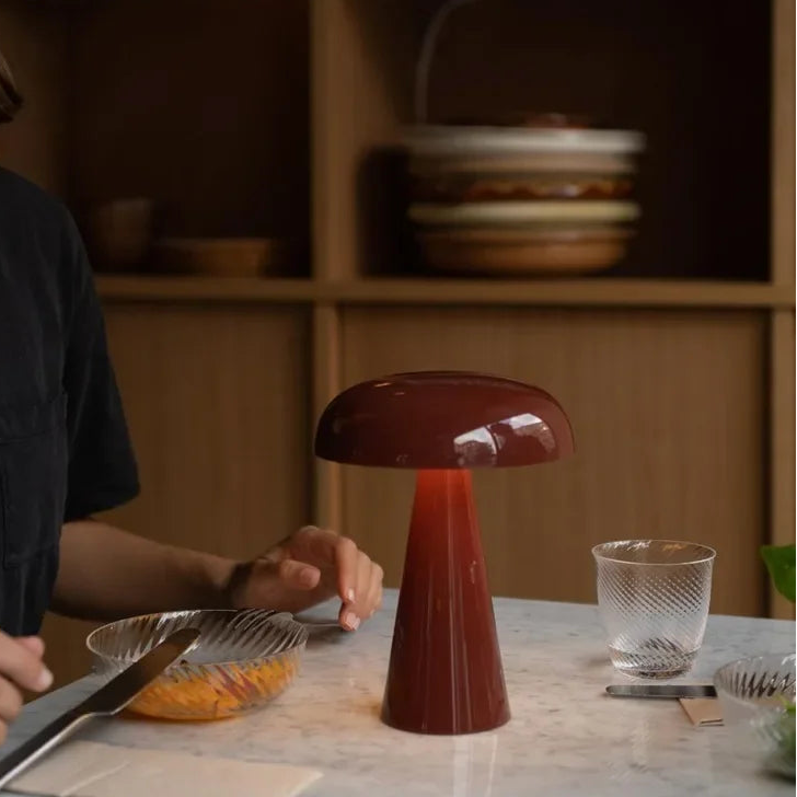 Red mushroom-shaped lamp on a table with a person and kitchen items in the background.