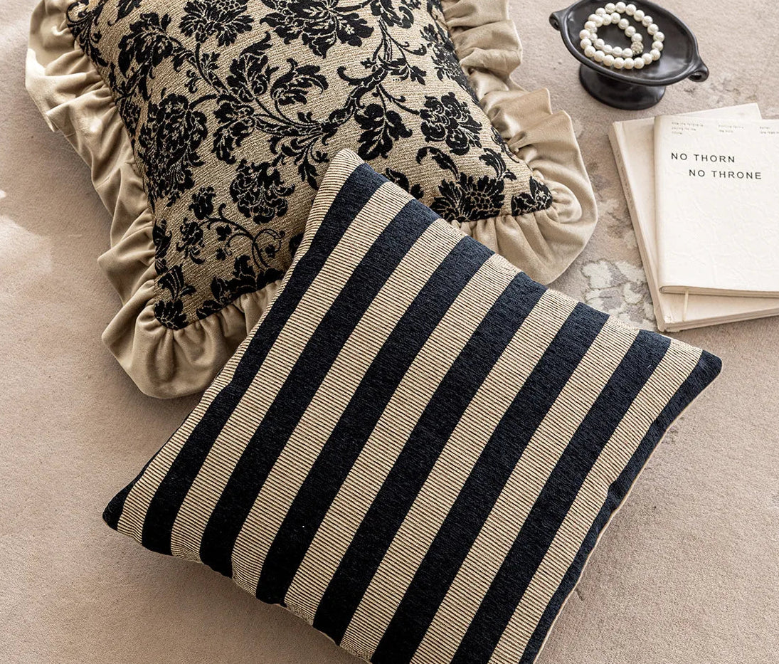 Two decorative pillows, one with black floral pattern and ruffled edge, the other with black and beige stripes, on a textured surface.