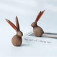 Two wooden rabbit figurines on a book titled 'The art of libraries' with a light gray background.
