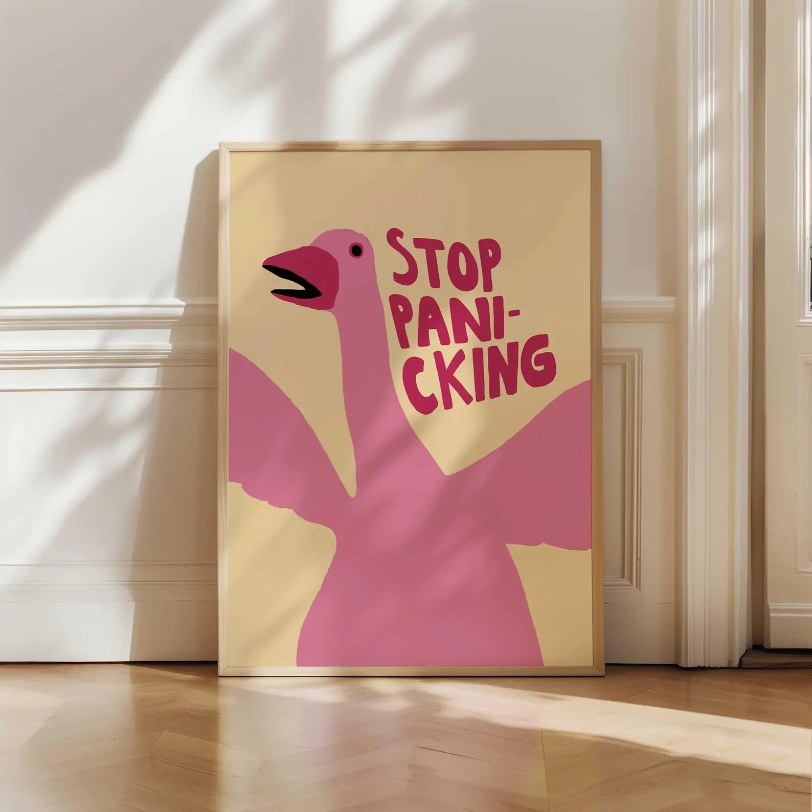 Framed artwork with a pink flamingo and text 'STOP PANIC-KING' on a beige background, leaning against a white wall.
