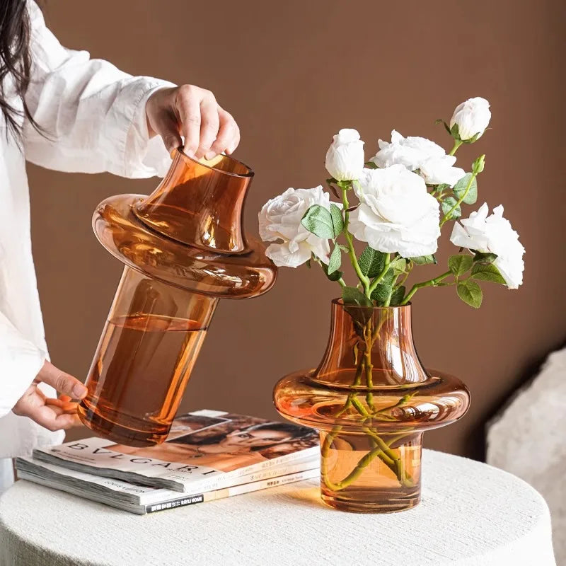 Person holding an amber-colored glass vase with a unique design, next to a matching vase with white flowers on a table.