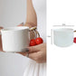White mug with cherry and gold handle held by a hand, with measurement details.