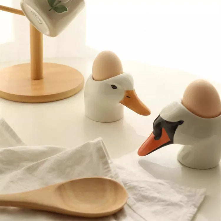 Two swan-shaped egg holders with eggs on a light surface with a wooden spoon and towel.