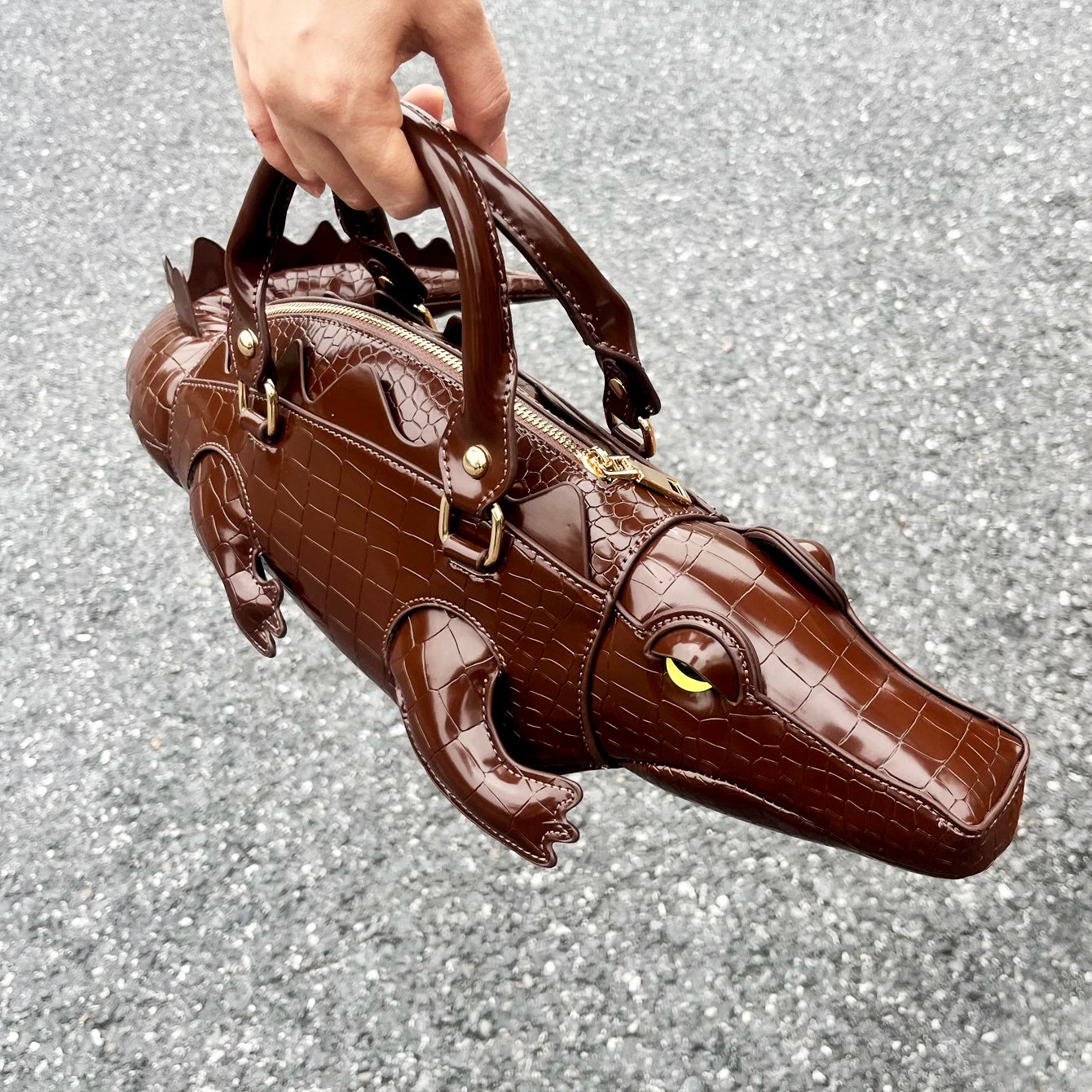 Brown alligator-shaped handbag held by a hand on a glittery gray background