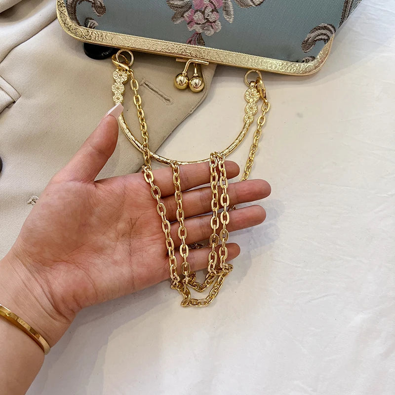 Gold chain necklace held by a hand with a beige bag and floral pattern in the background