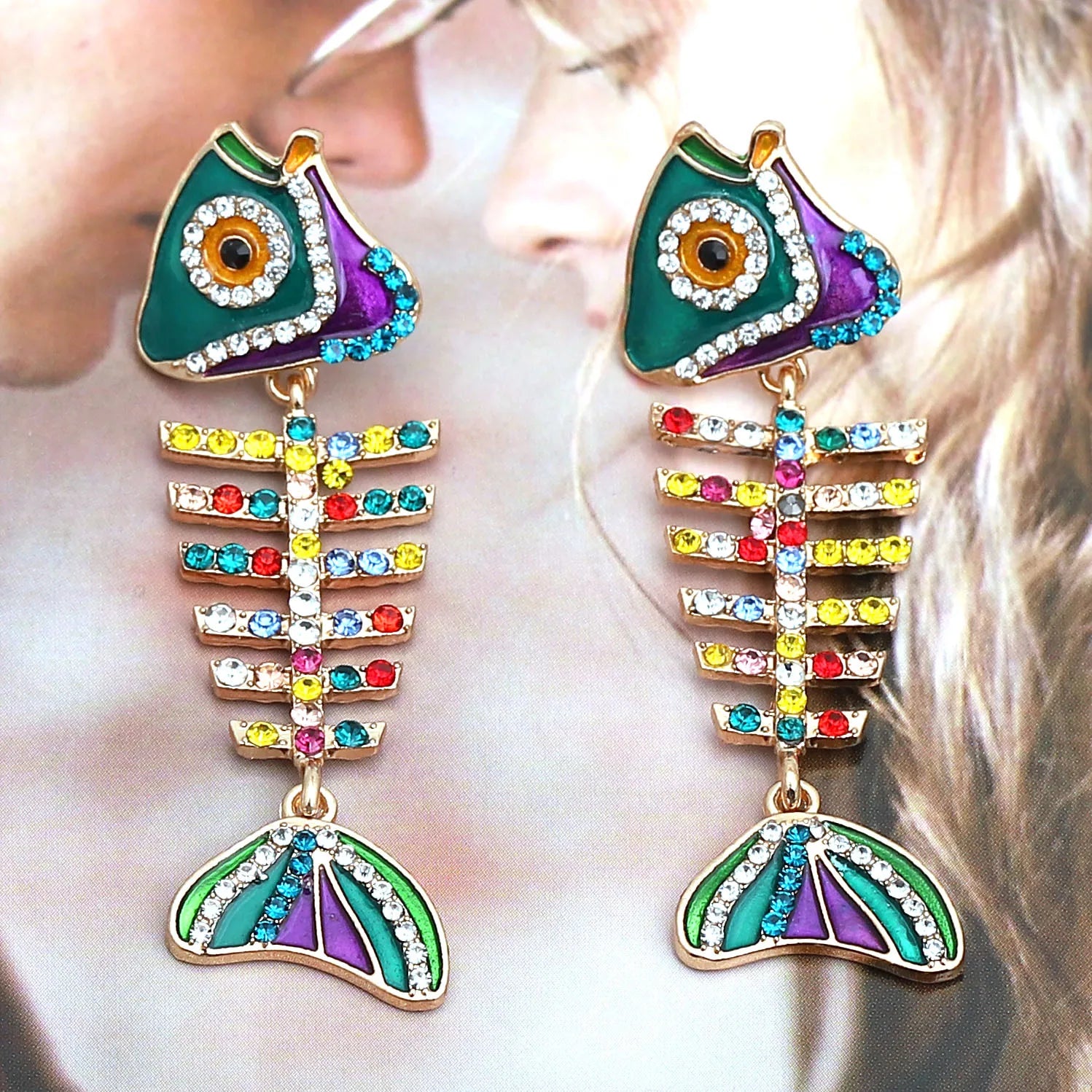 Colorful fish bone-shaped earrings with rhinestones worn by a person.