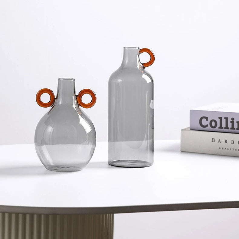 Two clear glass vases with orange handles on a white surface.
