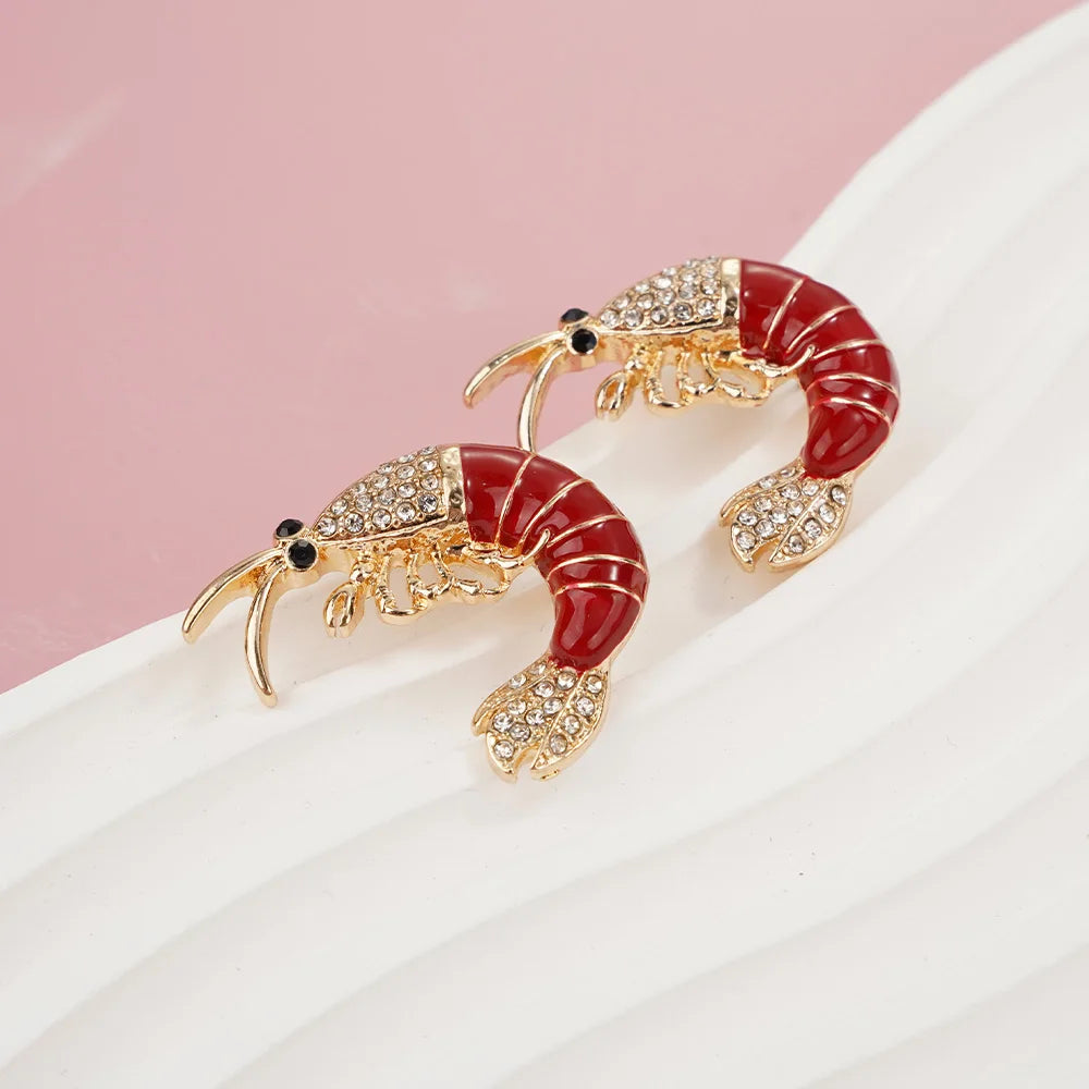 Shrimp-shaped earrings with red bodies and gold accents on a white surface.