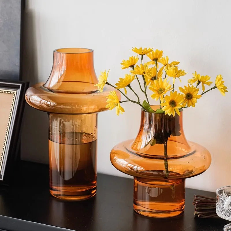 Two orange glass vases with one containing yellow flowers on a dark surface.