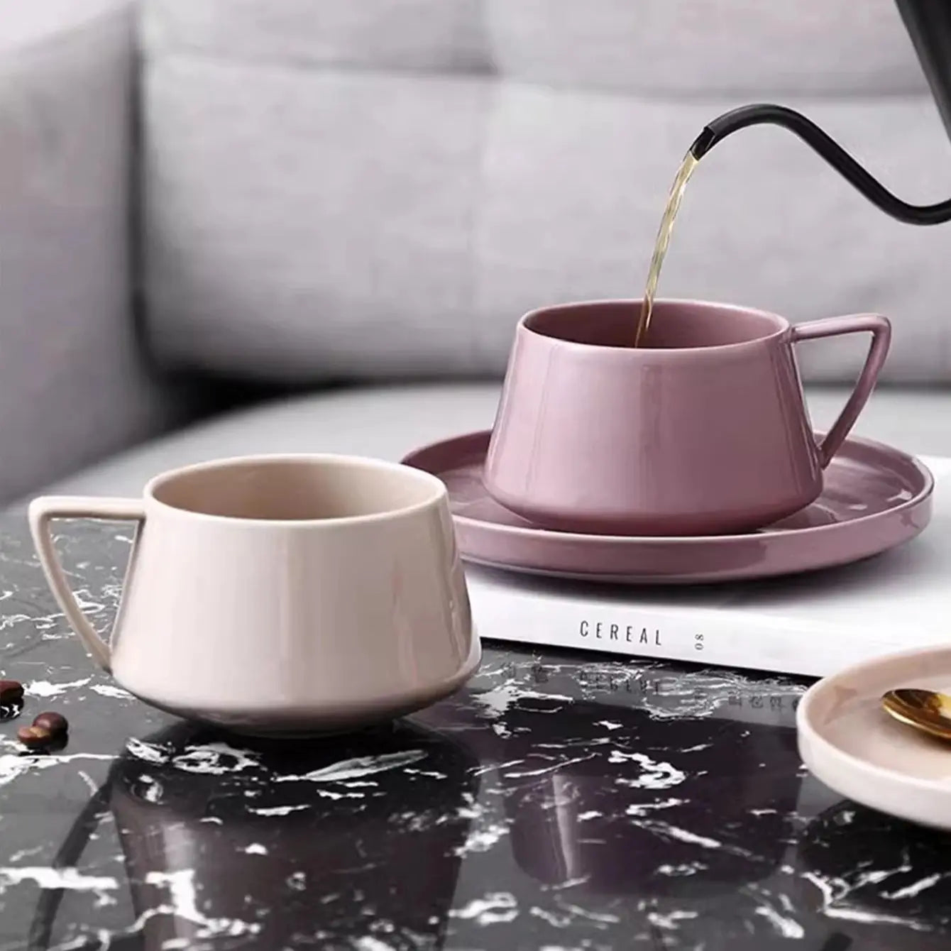 Pink mug being filled with tea on a marble surface with a white mug and plate.