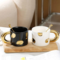 Two ceramic mugs with gold handles and spoons on a wooden tray.