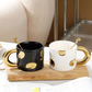 Two ceramic mugs with gold handles and spoons on a wooden tray.