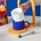 Colorful mugs with a yellow stand on a white surface, with a colorful box in the background.