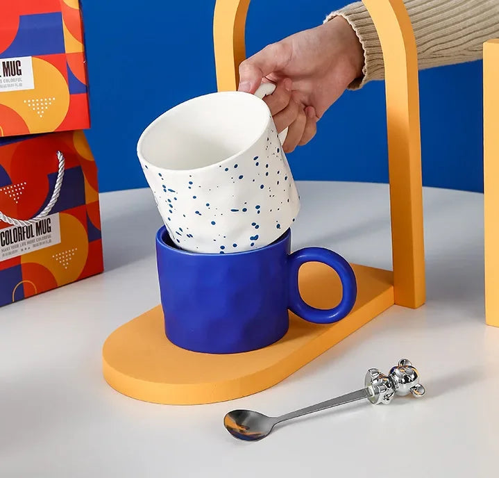 Colorful mugs with a yellow stand on a white surface, with a colorful box in the background.