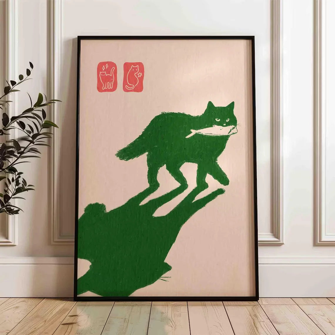 Framed artwork of a green cat with a fish on a beige background, displayed in a room.