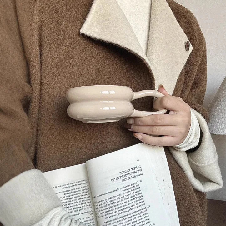 Person holding a beige ceramic mug with a book open in their lap, wearing a brown coat and white gloves.
