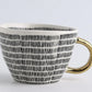 Ceramic mug with black pattern and gold handle on a light gray background, featuring the brand 'Fantasy'.