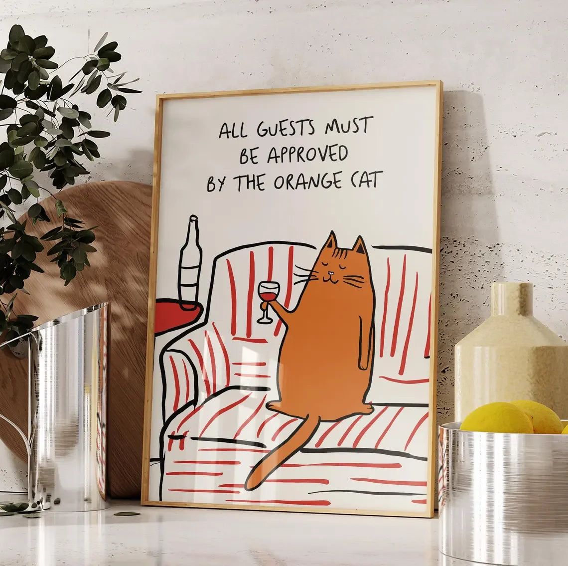 Framed artwork with a cat holding a glass of wine and text 'All guests must be approved by the orange cat' on a white wall.